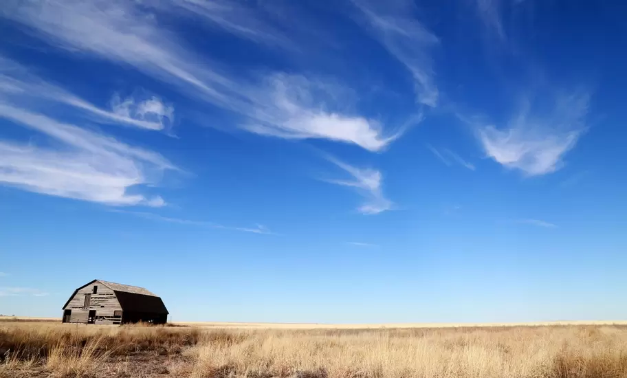 Weathered wooden barn alone on dry grassland in Oklahoma under a wide blue sky with wispy clouds