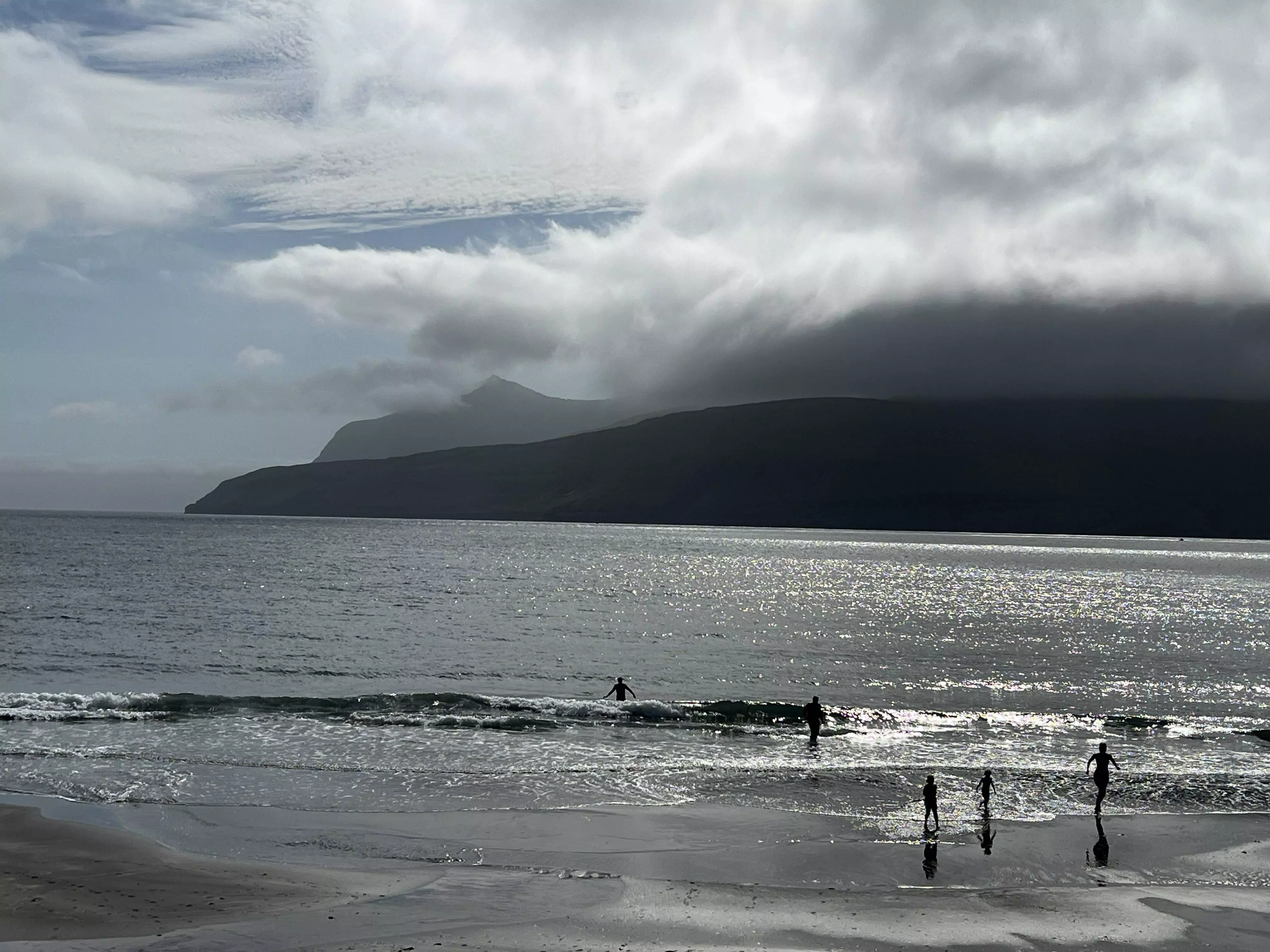 A family plays in the waves the water glistening from the sun, mountains are contrasted in the backgroud.