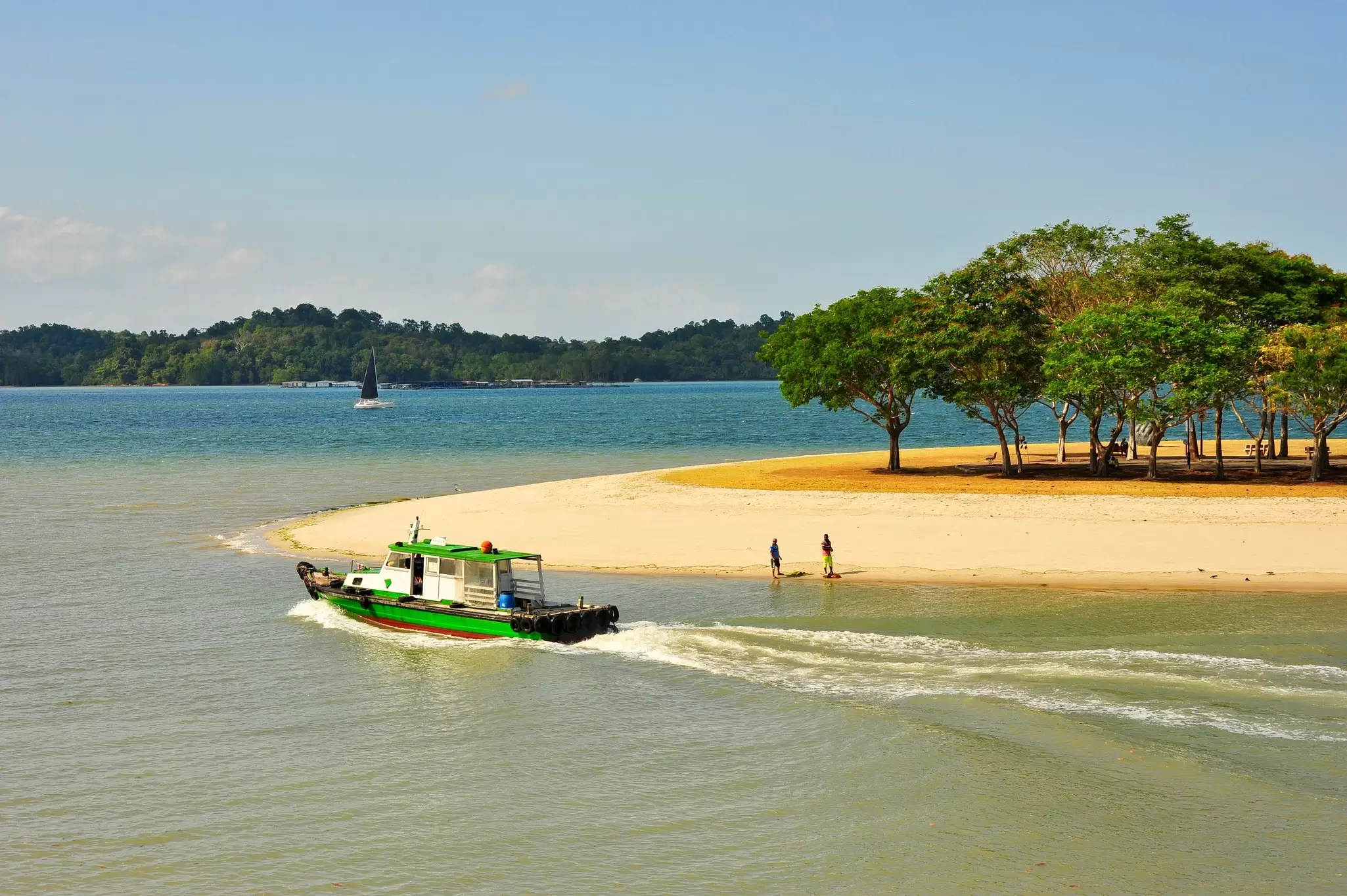 A sandy stretch of beach with a shady area under trees. Two people stand near the shoreline as a small boat and a yacht sail nearby.