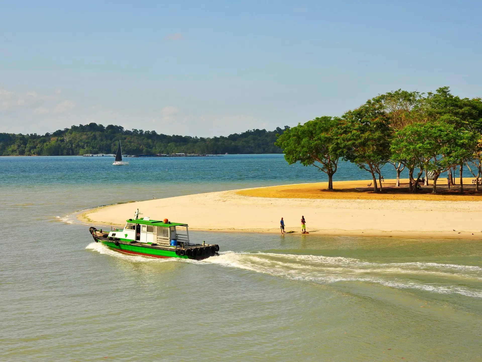 A sandy stretch of beach with a shady area under trees. Two people stand near the shoreline as a small boat and a yacht sail nearby.