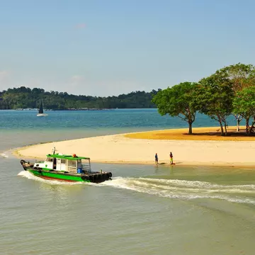 A sandy stretch of beach with a shady area under trees. Two people stand near the shoreline as a small boat and a yacht sail nearby.