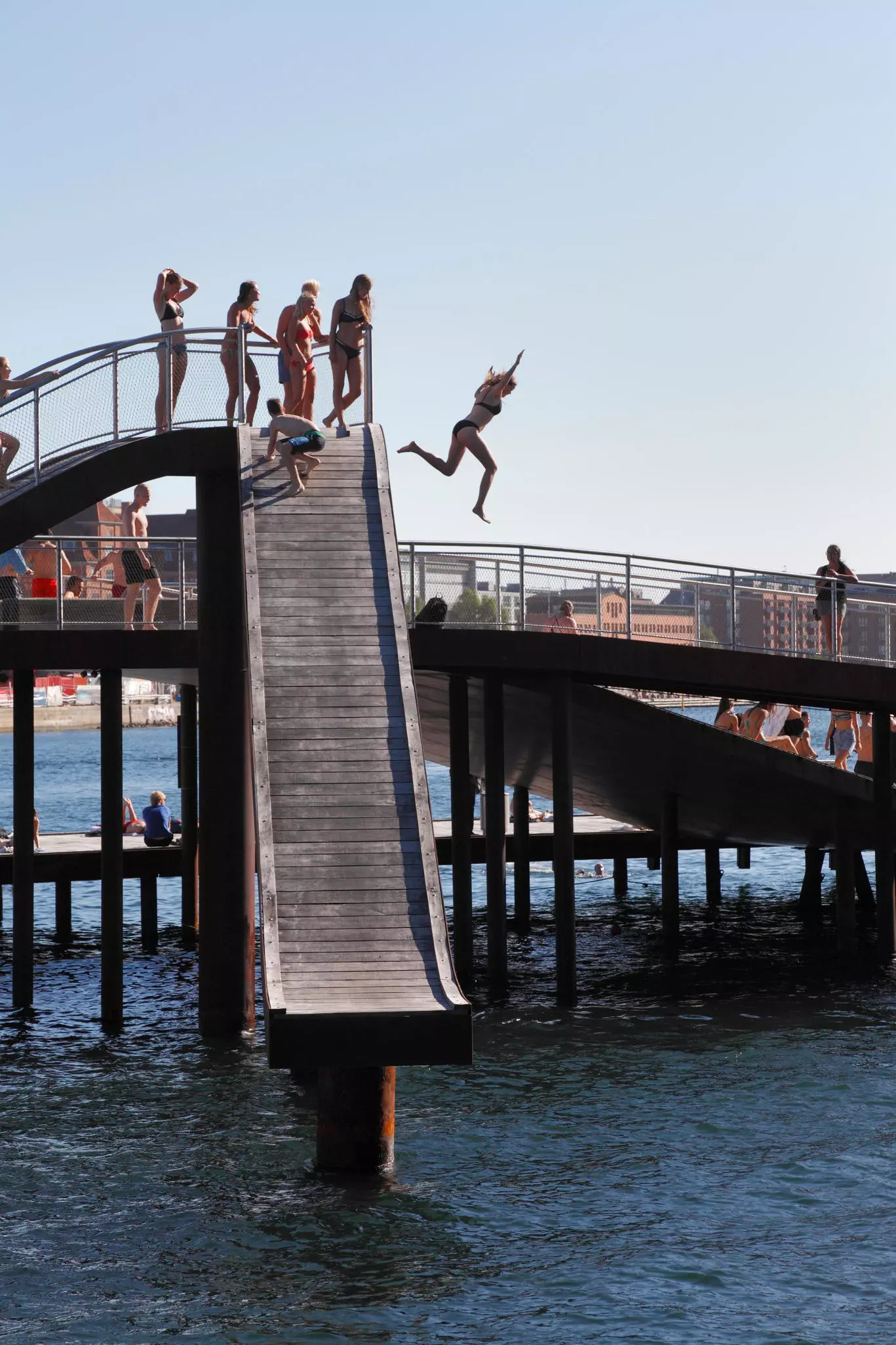People are standing at the top of a high wooden slide structure, while a woman jumps off the side into the water.