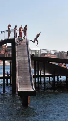 People are standing at the top of a high wooden slide structure, while a woman jumps off the side into the water.