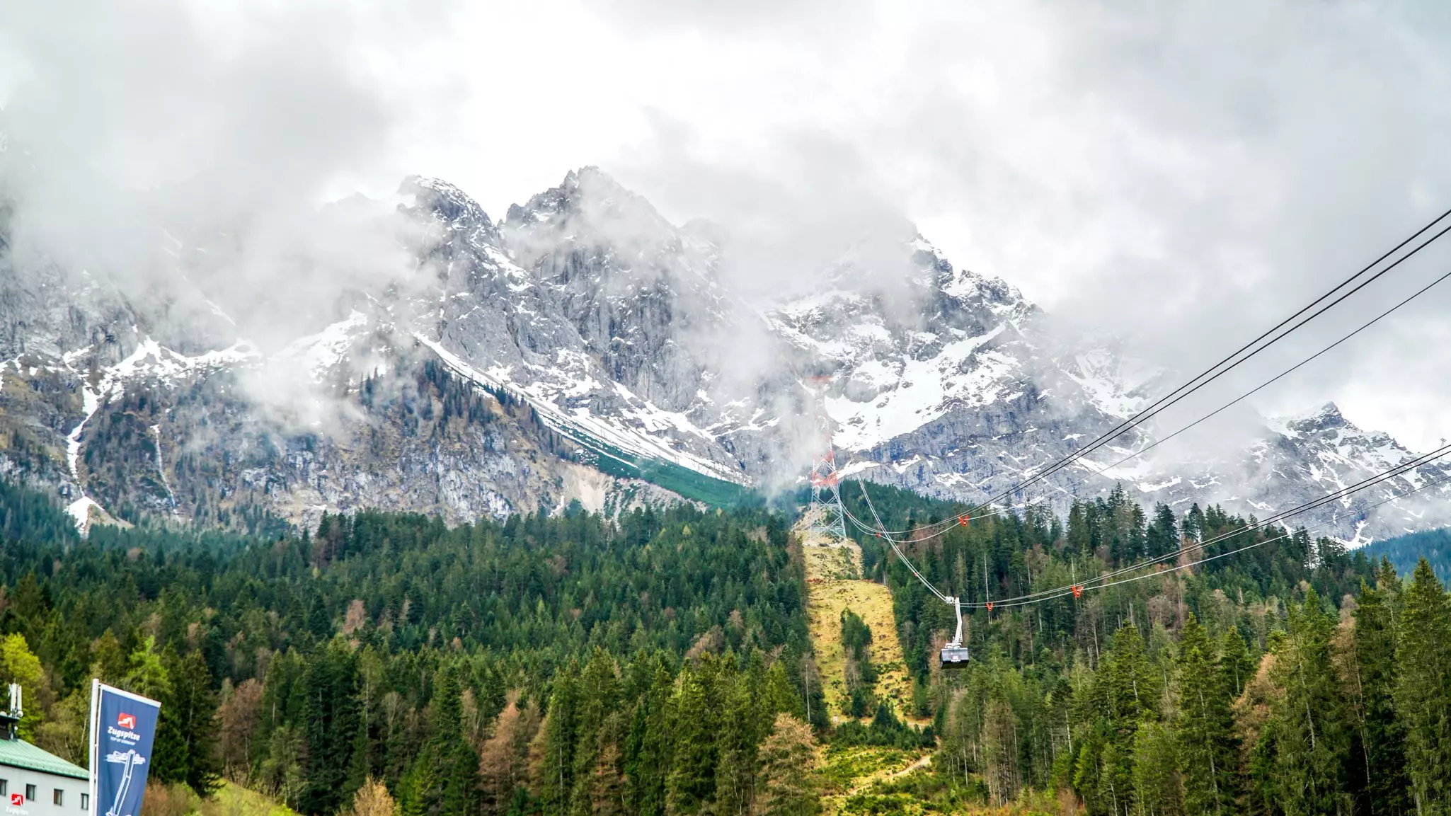 A cable car on a long span on the side of a snow-capped mountain shrouded in clouds.