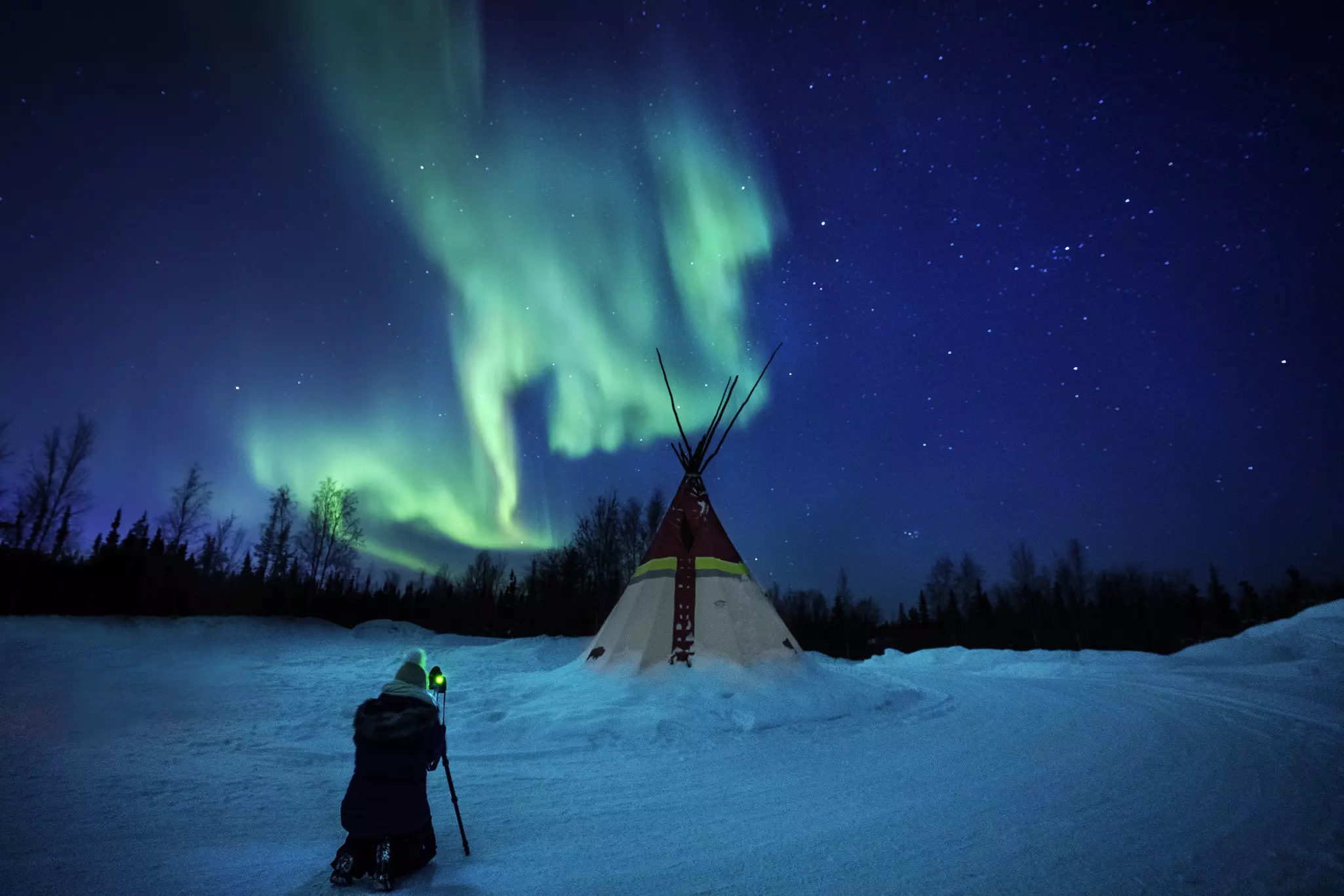 GettyA photographer in winter clothing takes a photo of a brightly lit traditional teepee under the green glow of the Northern Lights in a snowy landscape