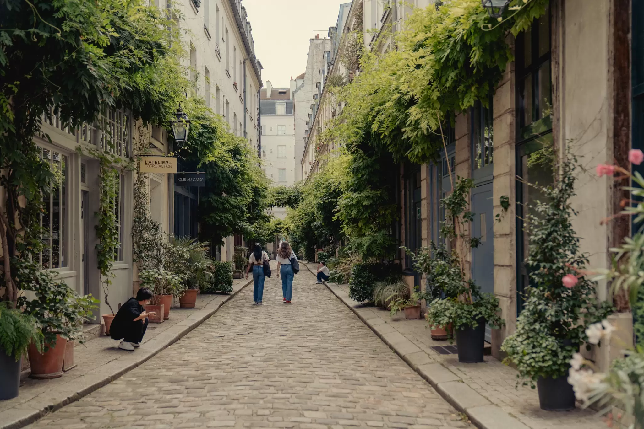 Two woman walk down a charming brick-lined street with classic French architecture and fern-like plants hanging over the doors