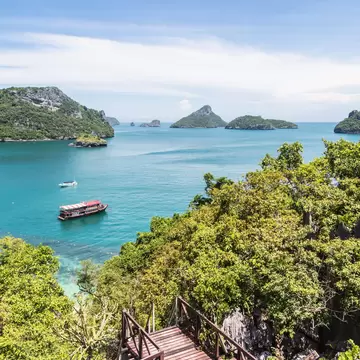 Wooden stairway through green scrub above a turquoise bay with moored boats and rocky forested islands, Ang Thong, Thailand.