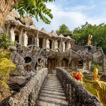 The exterior of a temple covered in stones and shells from floor to ceiling, surrounded by trees.
