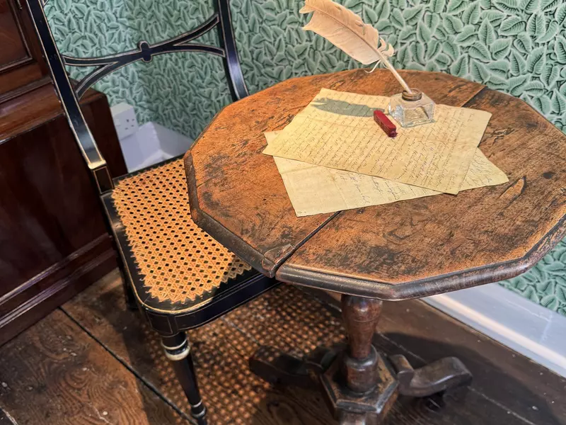 Sheets of paper and a quill on an octagonal dark wood table and a wood chair with a woven seat.