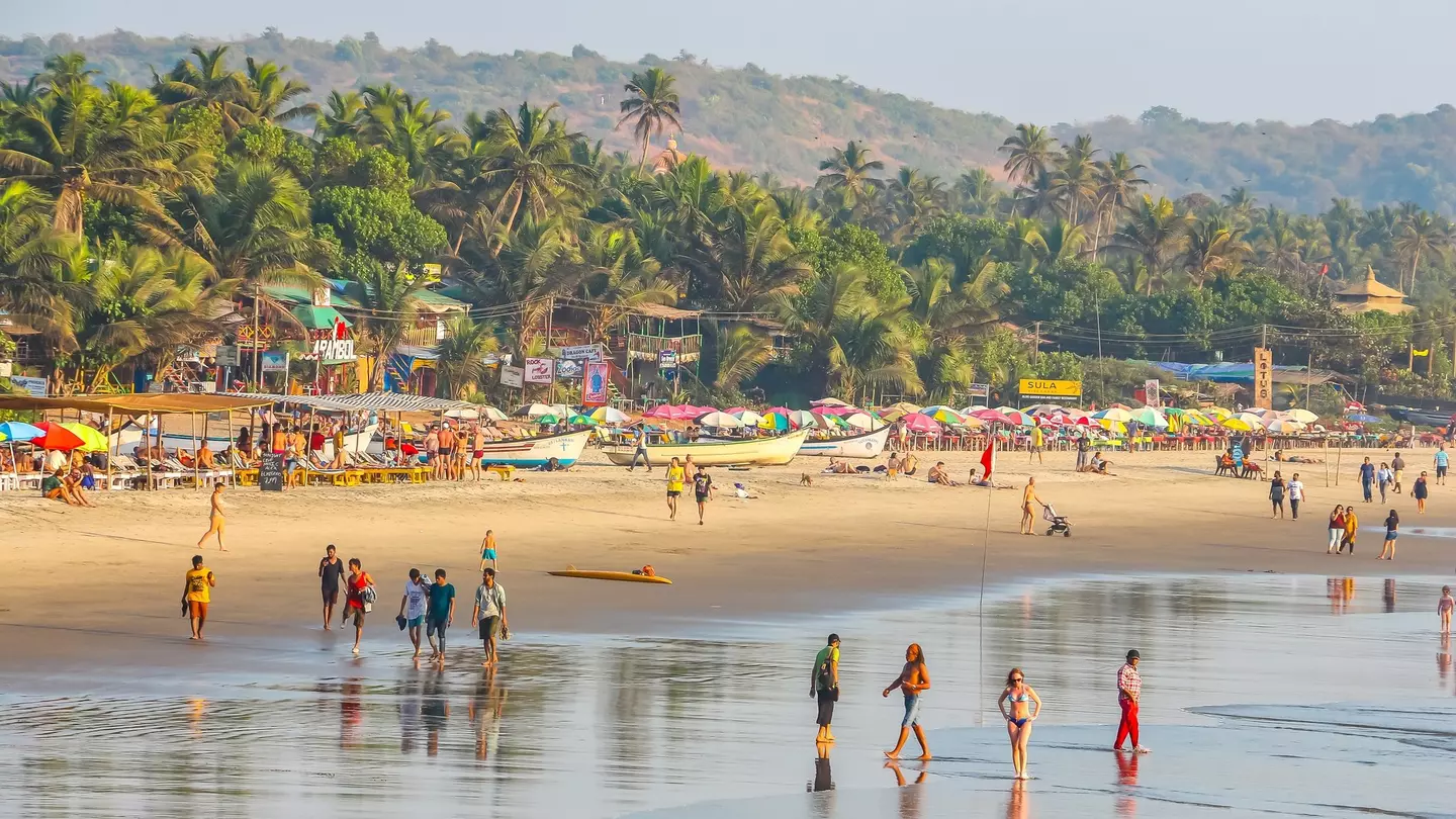A wide view of people walking in the surf of a wide tropical beach. Palm trees line the shore beyond boats and shacks on the beach.