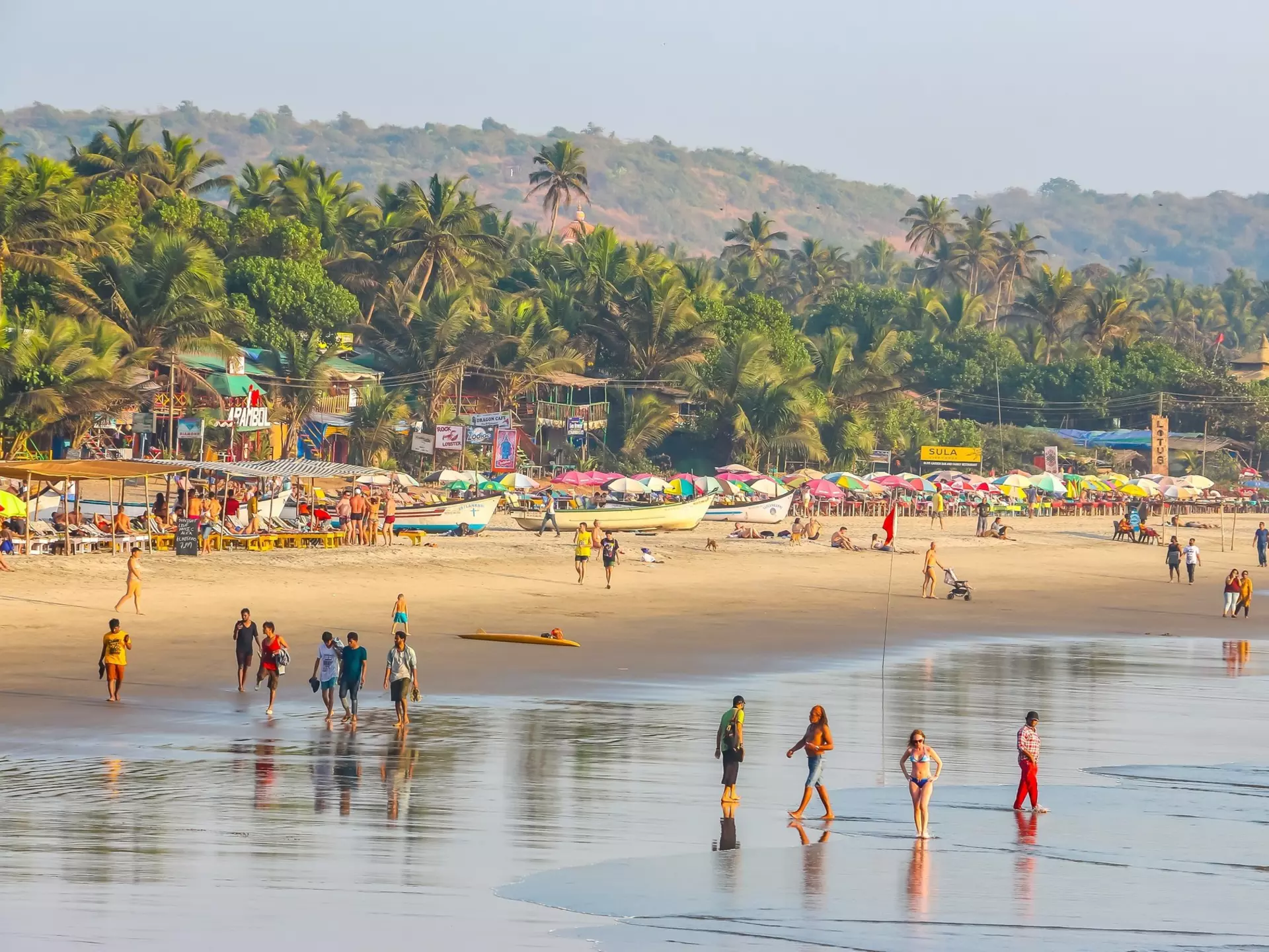 A wide view of people walking in the surf of a wide tropical beach. Palm trees line the shore beyond boats and shacks on the beach.