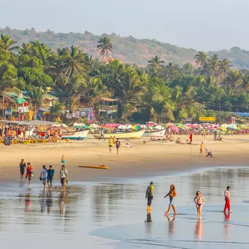 A wide view of people walking in the surf of a wide tropical beach. Palm trees line the shore beyond boats and shacks on the beach.