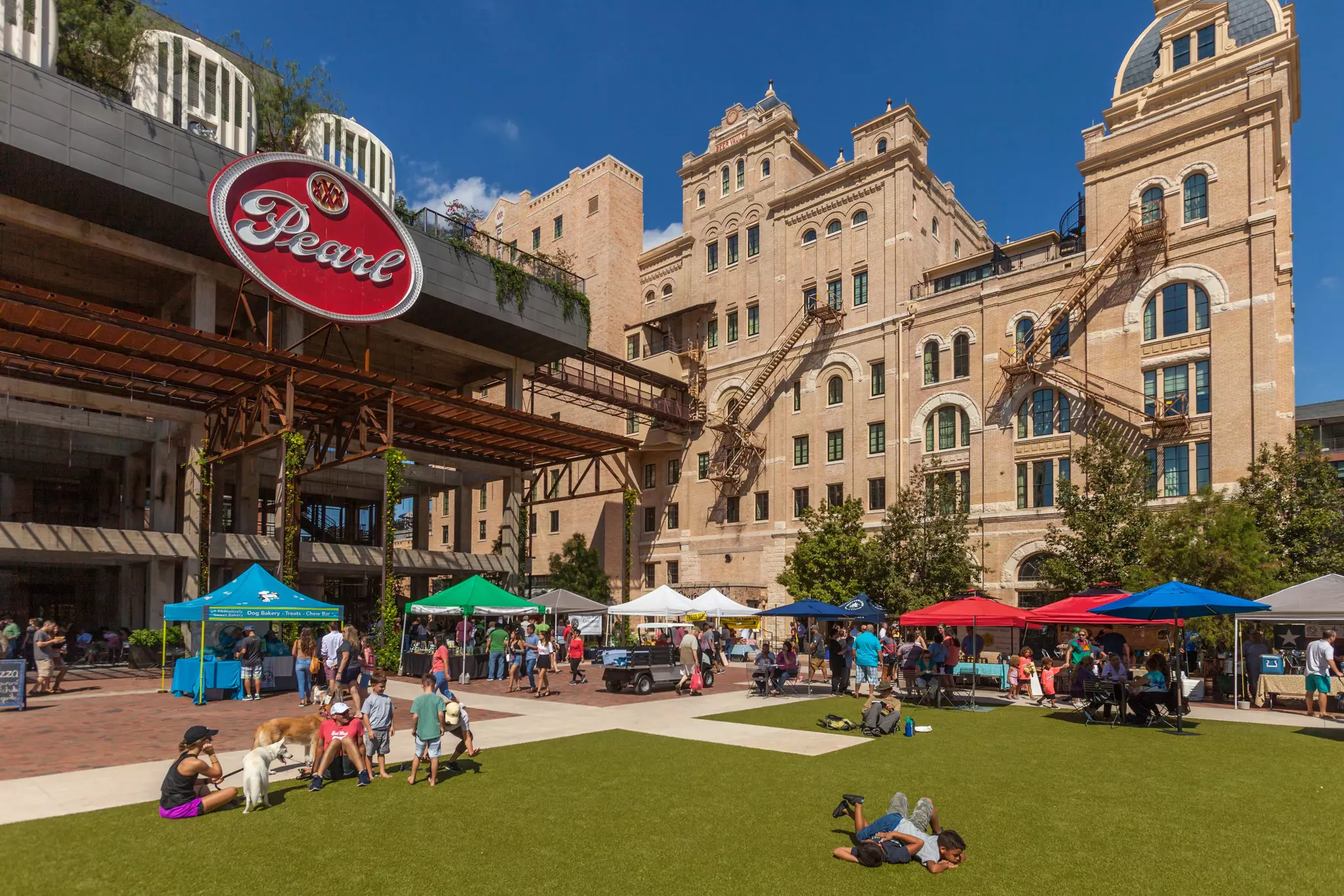 People sitting on a grass lawn and standing near tents with vendors; to the left is a building with a red sign reading "Pearl."