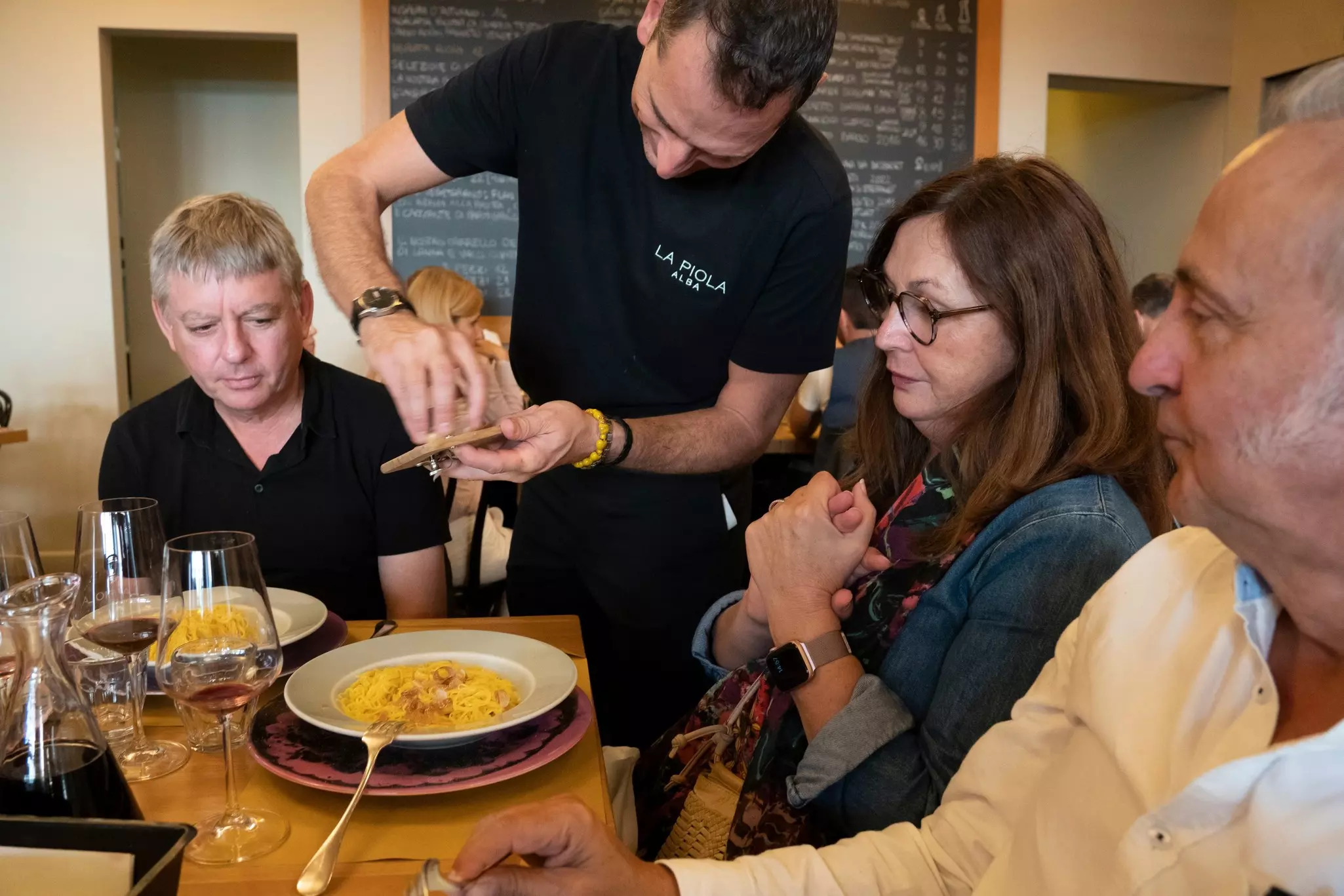 A waiter at a restaurant grates a truffle onto a diner’s order of pasta. Two other diners share her table.