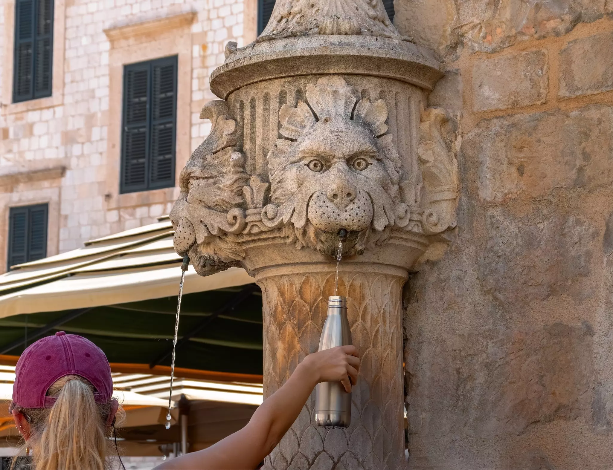 A person holding up a metal water bottle to a stream of water coming from a fountain with carvings.