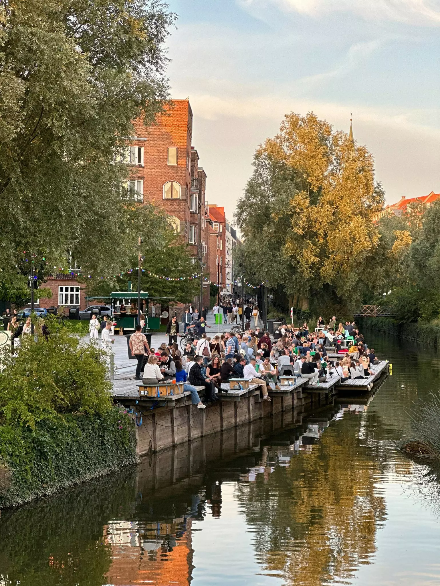 People are sat out on decking with drinks near a canal running through a town as the evening light of a summer's day begins to fade.