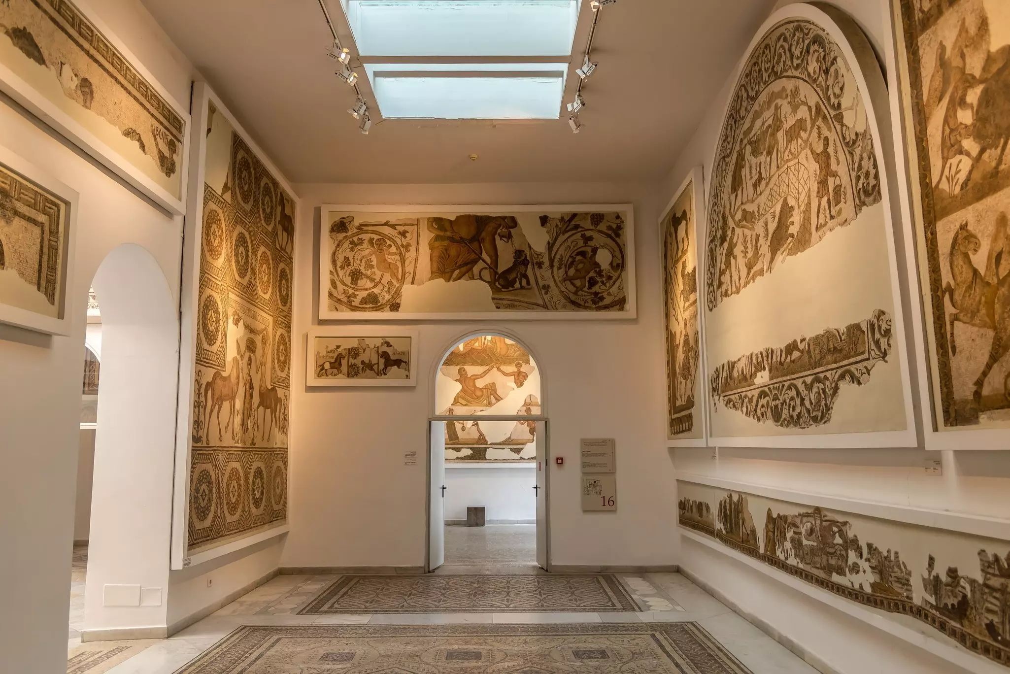 A room in a museum displaying mosaics of animals, people and scenes from daily ancient Roman life that have been pieced together on the walls and floors. Some parts are missing.