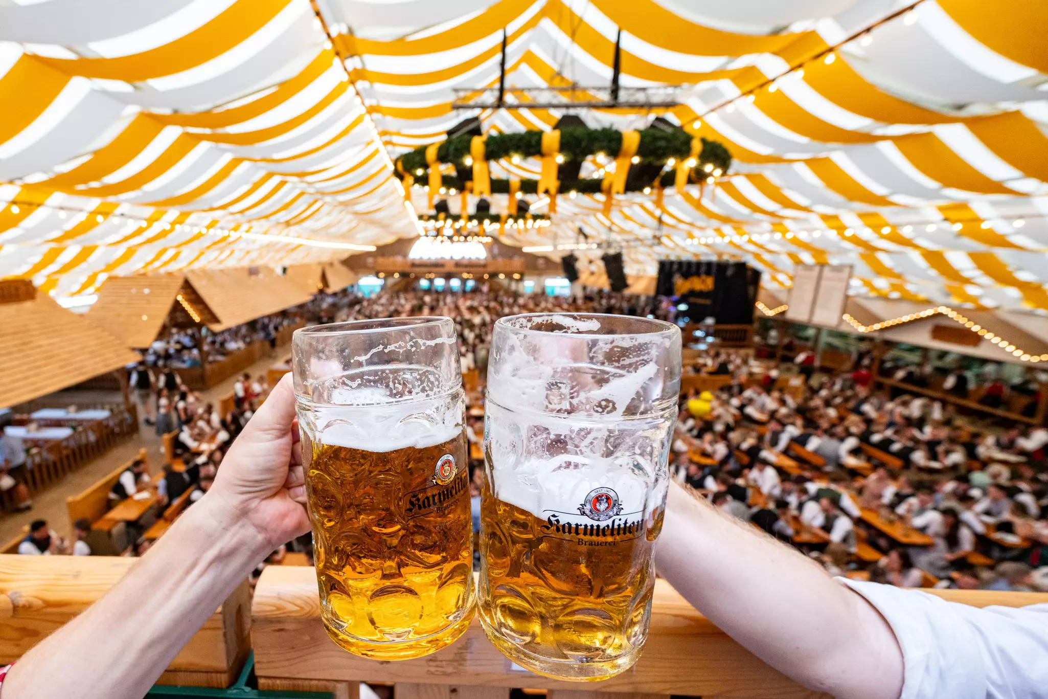 Two arms holding large glass steins of beer toast under a tent with a large seating area below.