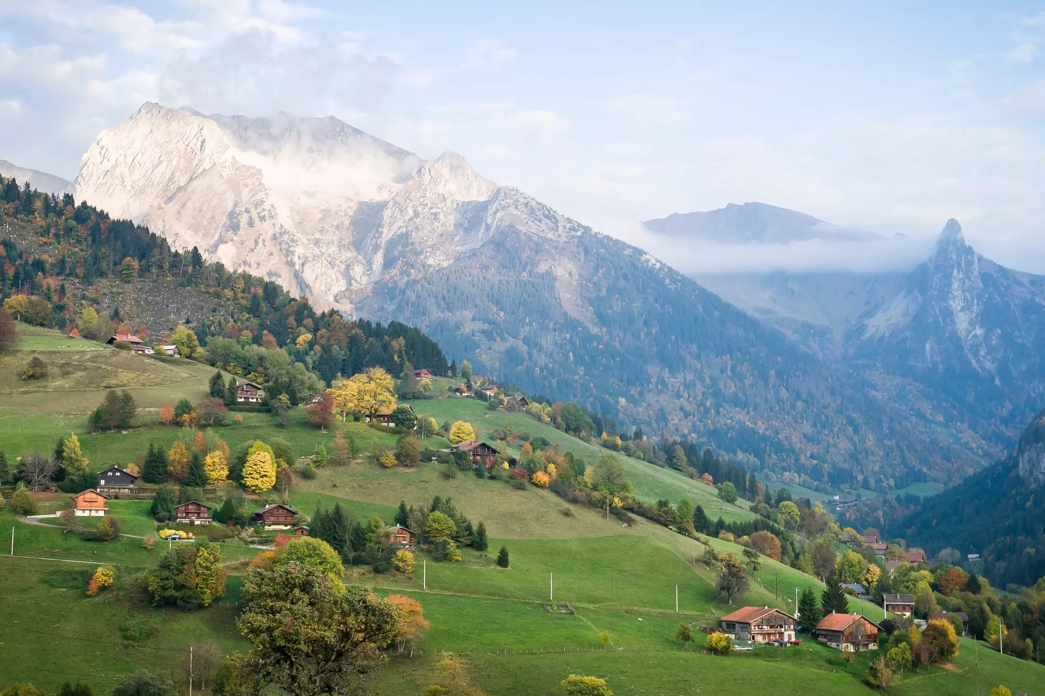 Wooden chalets in a mountainous region surrounded by green grass and trees as low cloud passes between mountain peaks.