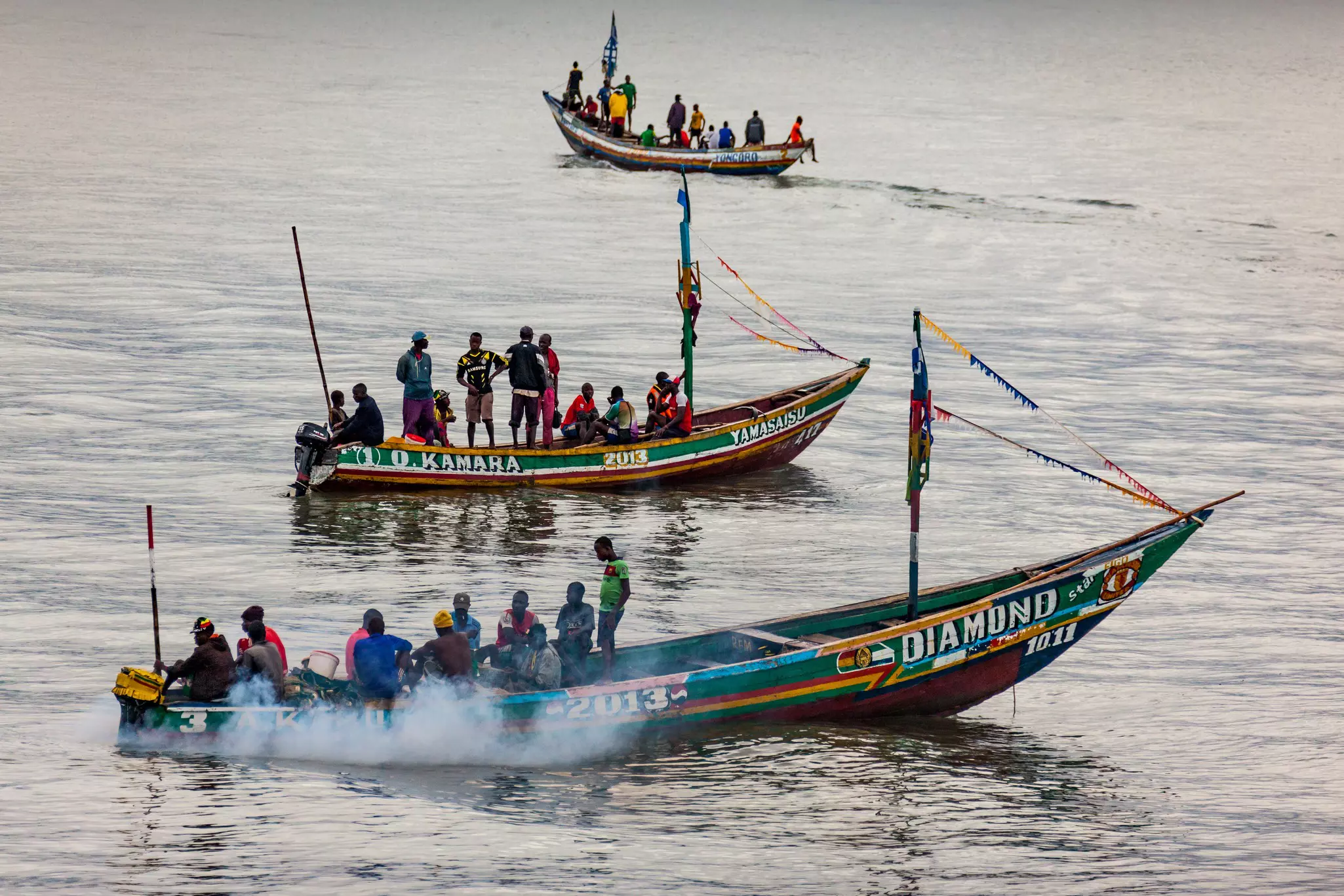 Three brightly painted boats carrying several fishermen each are pictured on the water.