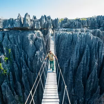 A narrow plank suspension bridge with wire handrails spanning a deep chasm between fields of jagged grey limestone pinnacles, a man in a pale shirt walking across it towards the camera.