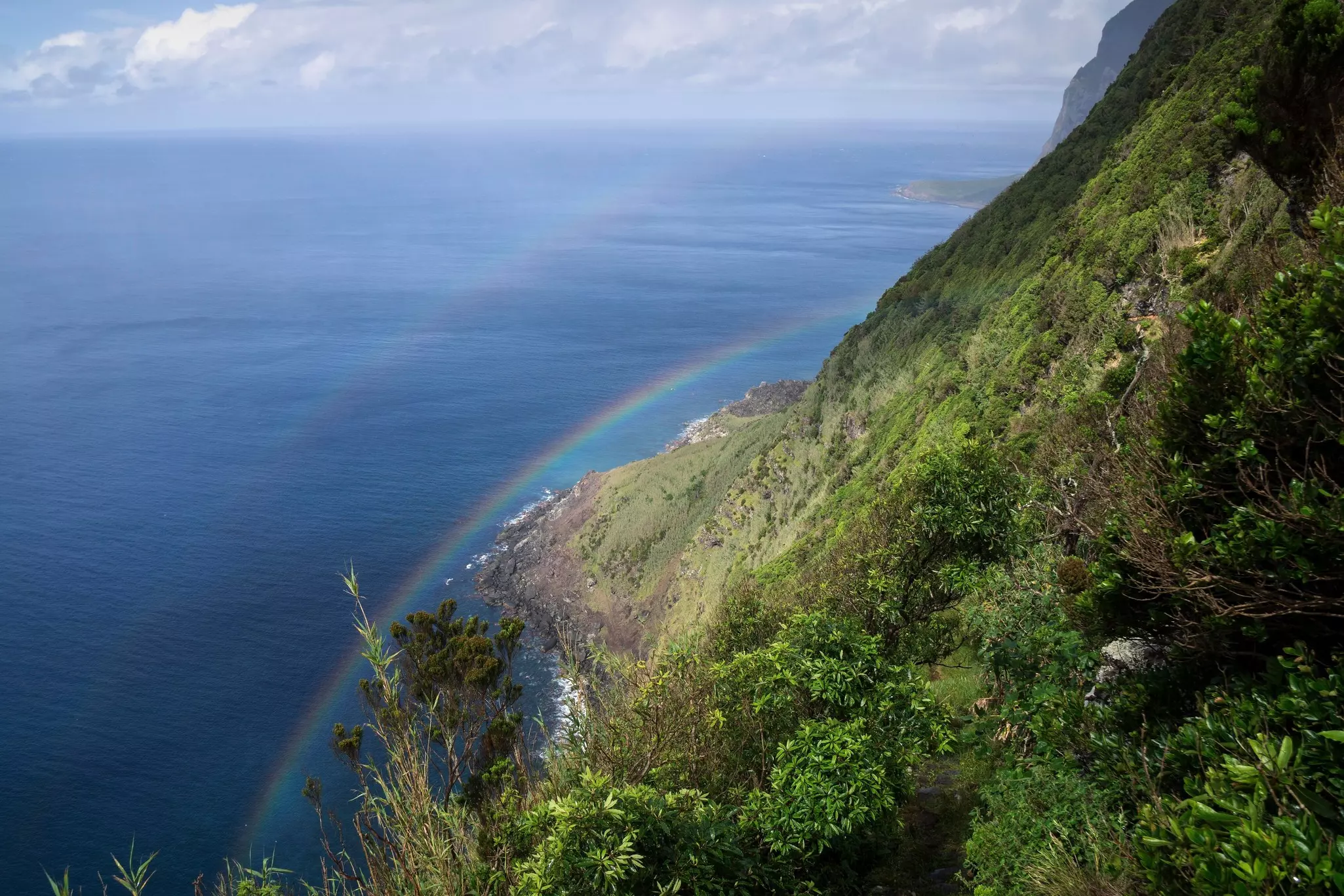 Steep coast with a double rainbow overlooking the ocean on a sunny day with some white clouds in sky.