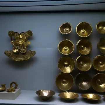 A display case holds a gold pectoral with a human face and rows of hammered gold bowls against a pale blue wall.