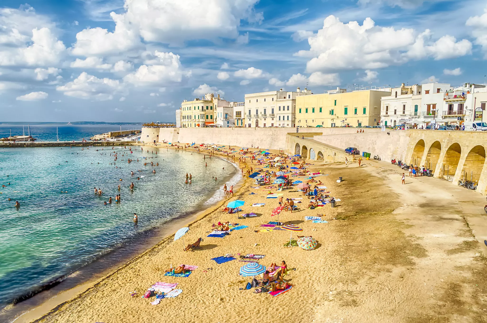 People relax on the beach on the waterfront in a town with a small harbor.