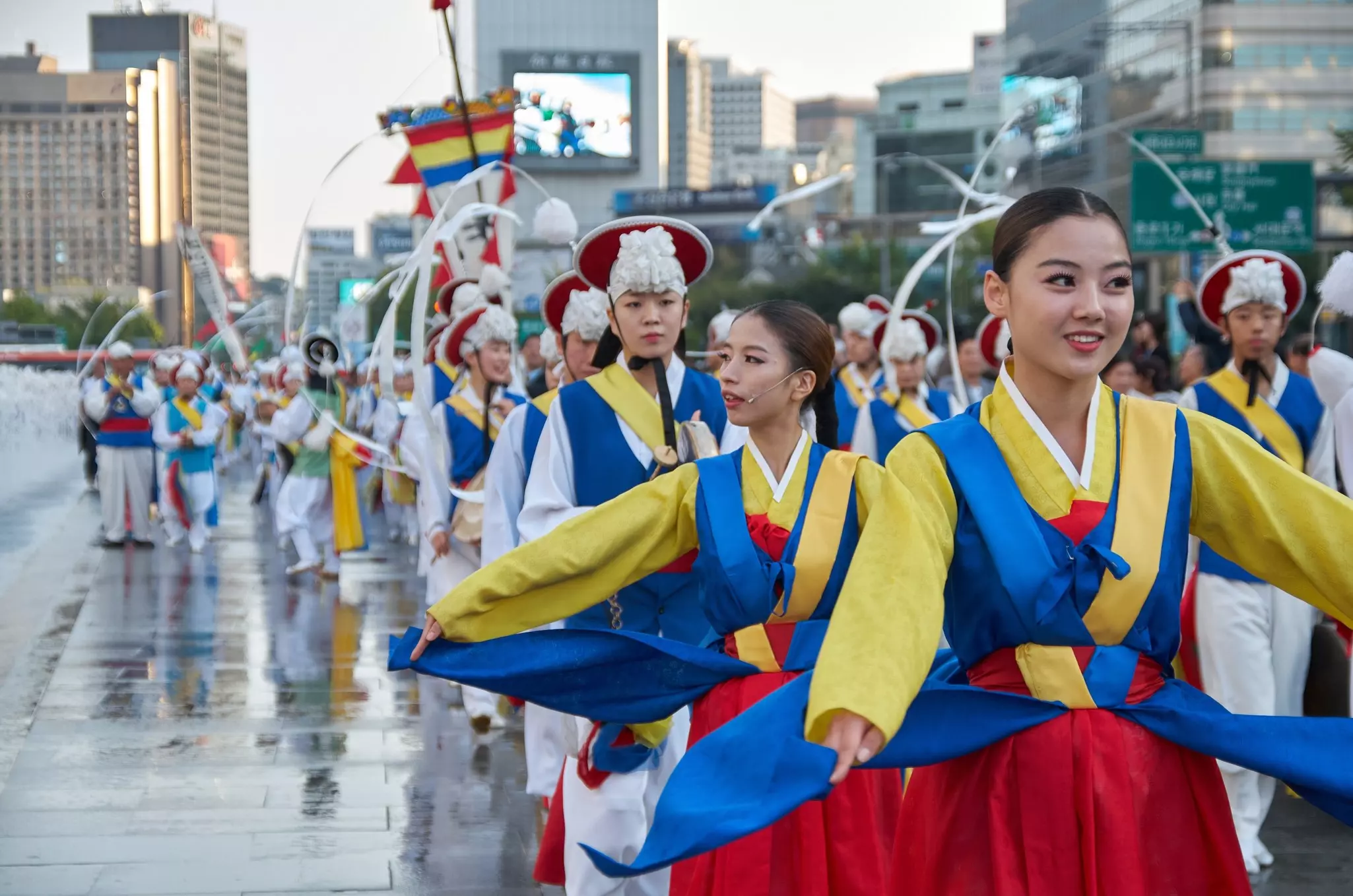 People in traditional robes perform in a procession on a rainy day in a city.