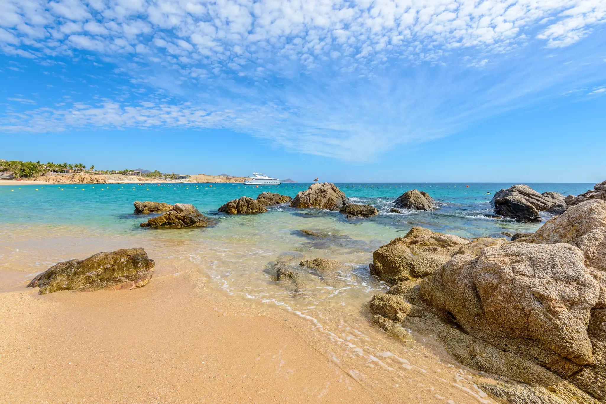 Clear shallow water off a sandy beach leads to rocks and blue sea beyond.