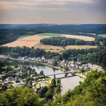 Hilltop view of a village along a river crossed by a bridge, with forests and golden fields beyond, in the Belgian Ardennes.