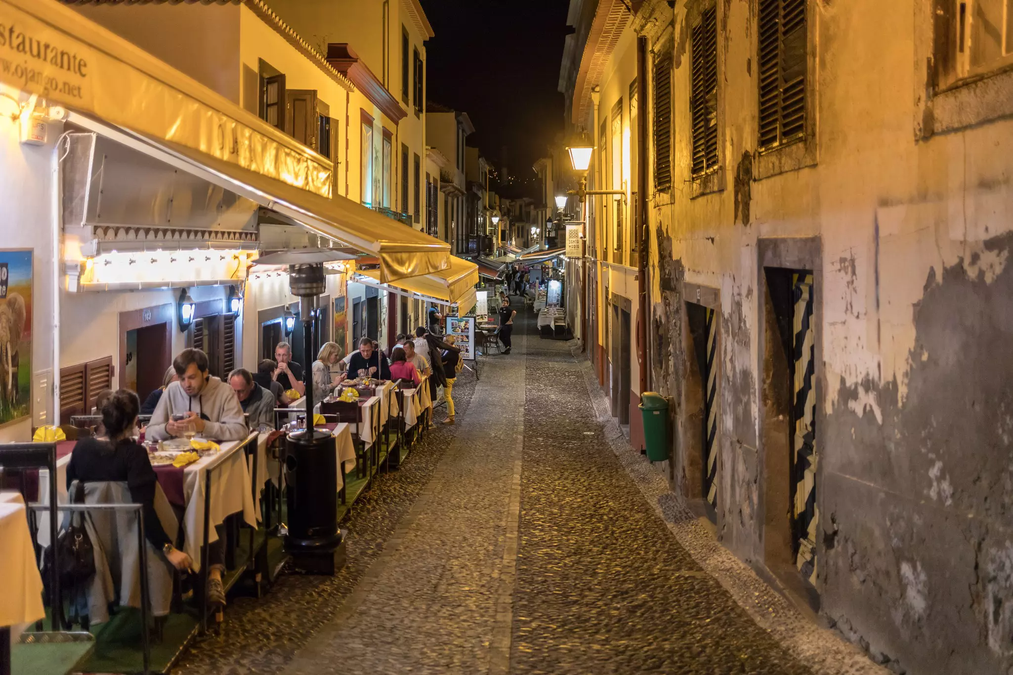People dine at night at outdoor tables along a narrow cobbled alley in a city.