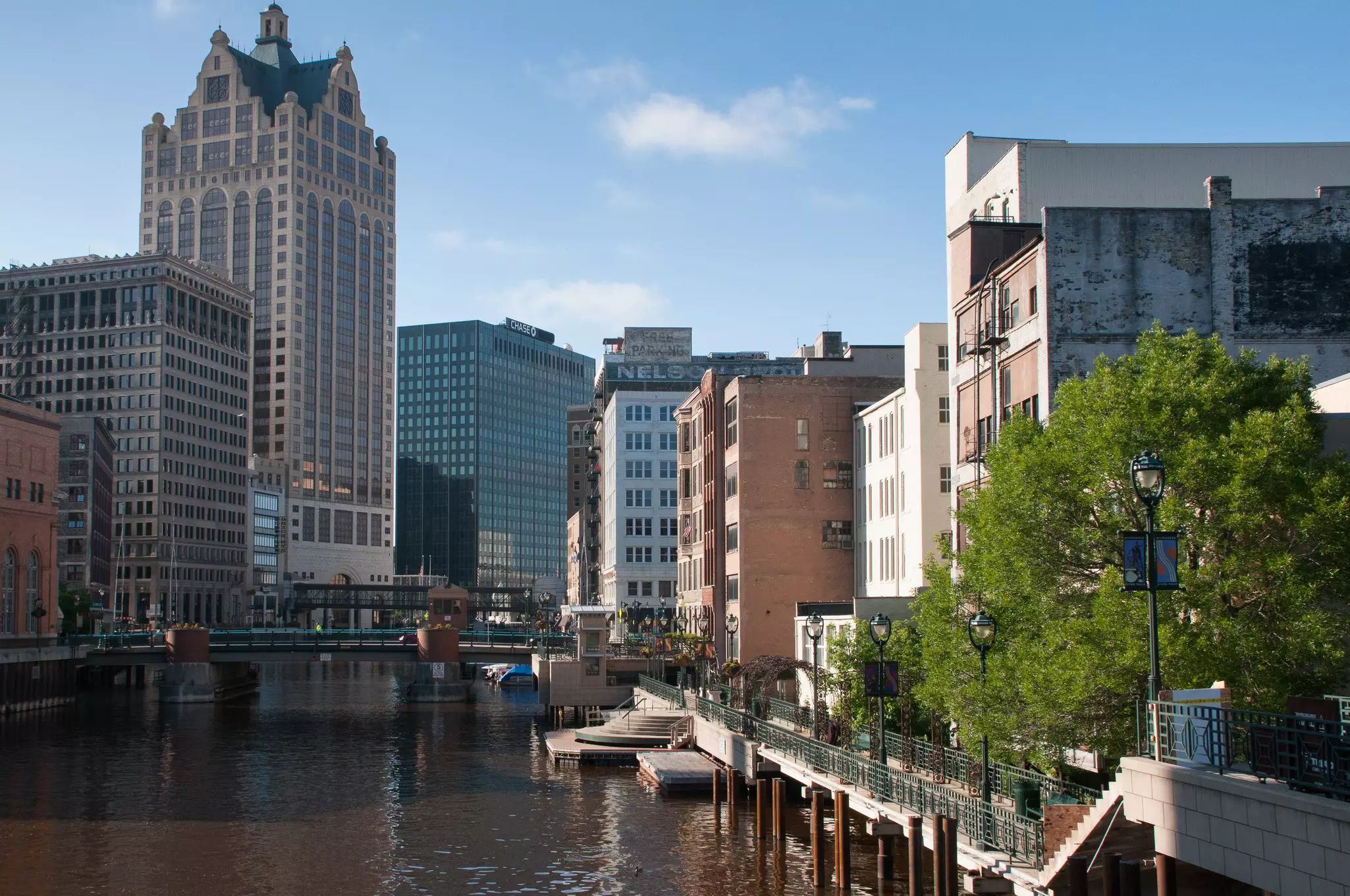 A pedestrian path leads along a river past large buildings in a city.