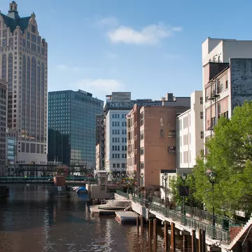 A pedestrian path leads along a river past large buildings in a city.