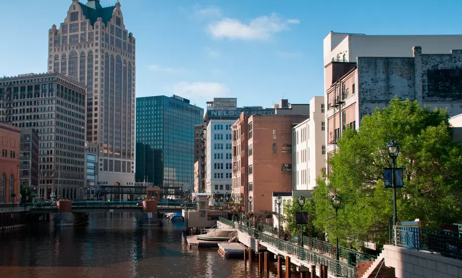 A pedestrian path leads along a river past large buildings in a city.