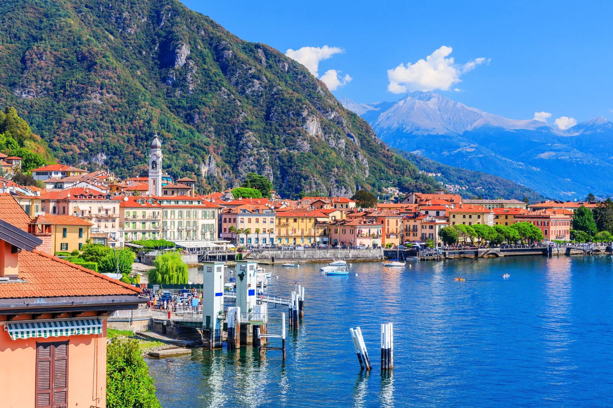 The shore of a town is pictured by a blue lake. A mountain rises behind the buildings of the town.