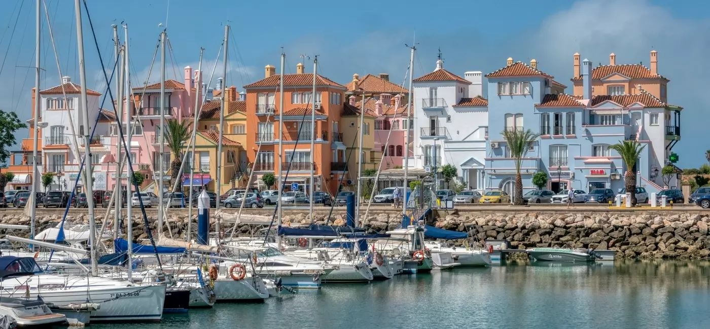 A row of pastel-colored four-storey buildings overlook a harbor where small boats are moored.