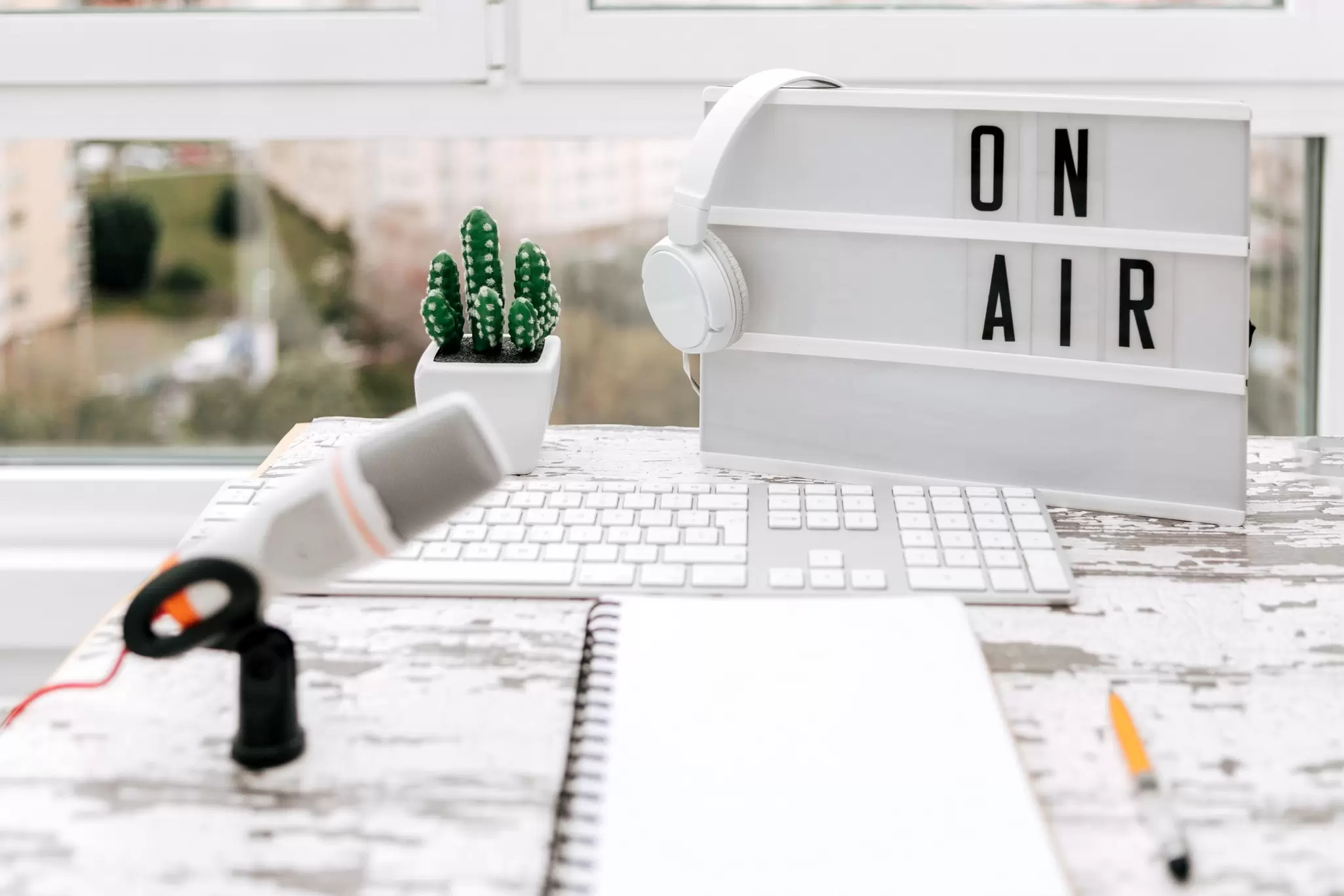 Podcast desk setup with microphone, white headphones, keyboard and an illuminated On Air lightbox sign by a window