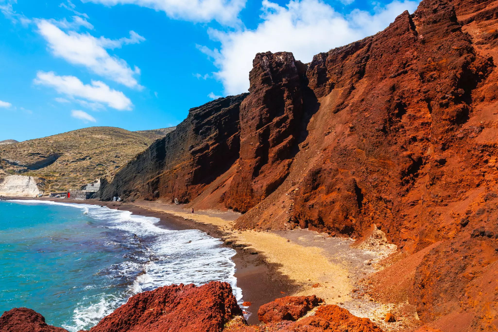 Red volcanic cliffs along a blue sea under light blue skies with thin clouds