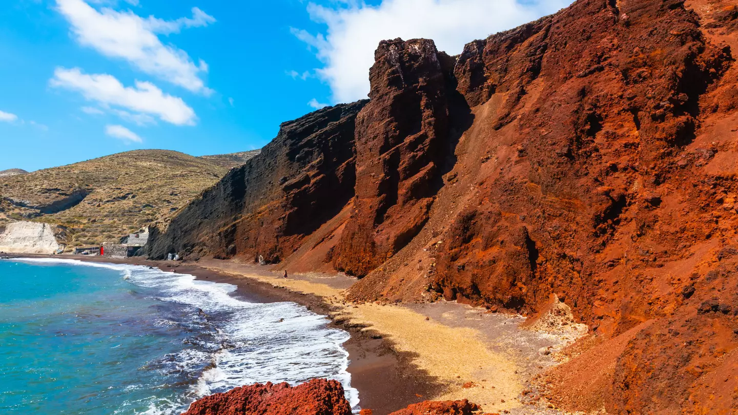 Red volcanic cliffs along a blue sea under light blue skies with thin clouds