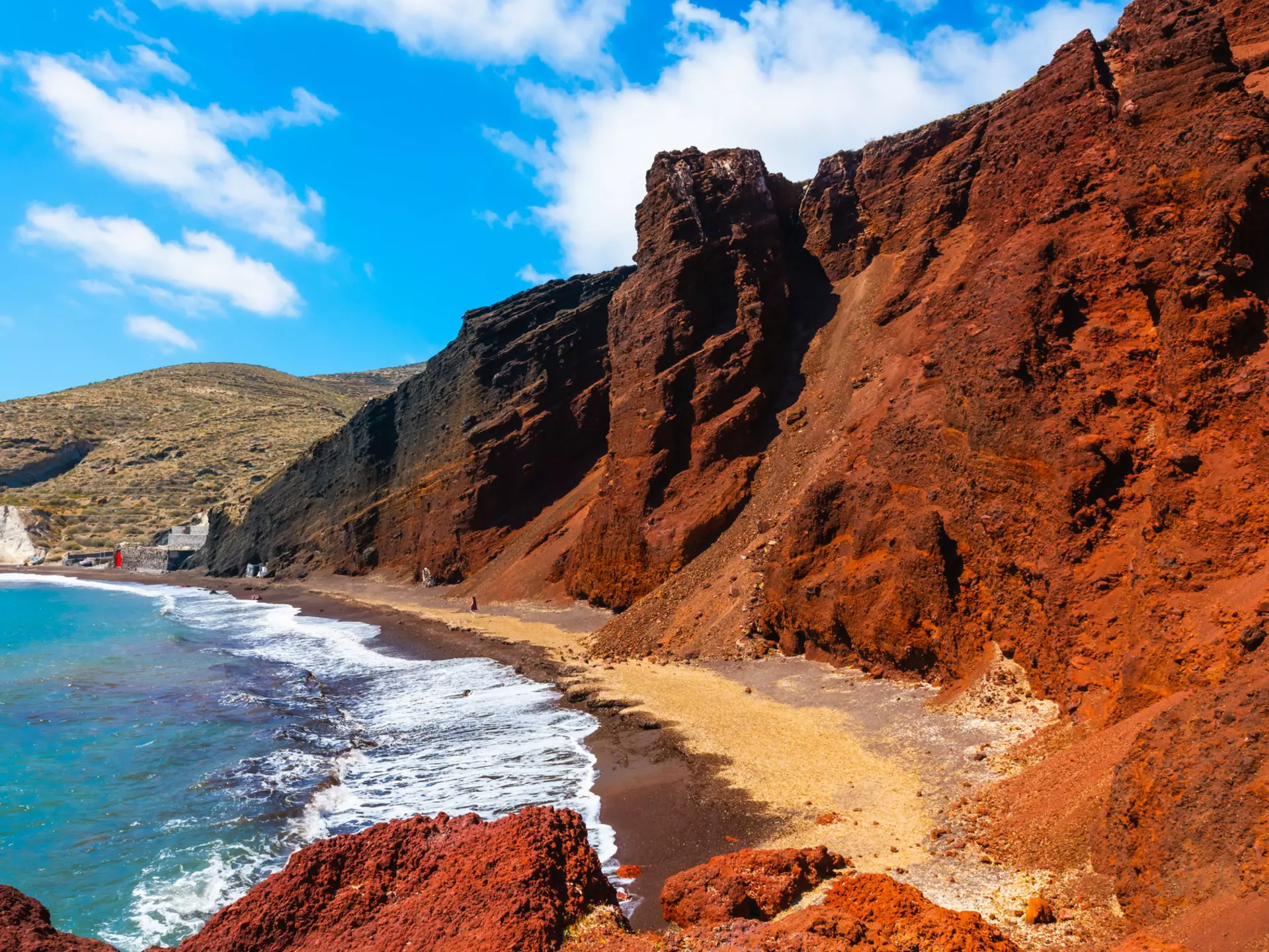 Red volcanic cliffs along a blue sea under light blue skies with thin clouds
