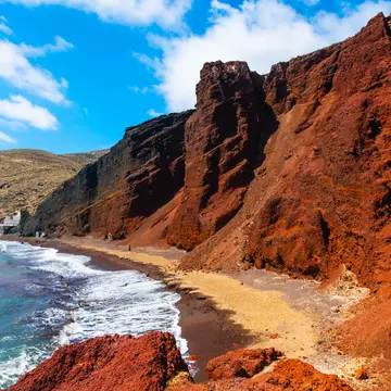 Red volcanic cliffs along a blue sea under light blue skies with thin clouds