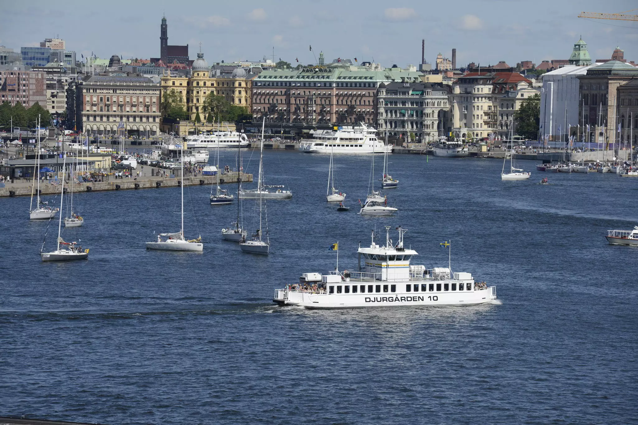 A ferry passes by moored sailboats in the busy harbor of a city.