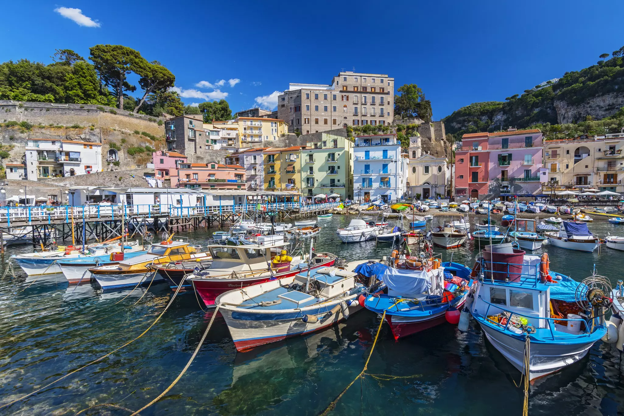 Image of small fishing boats in Sorrento's Marina Grande harbor with colorful buildings and trees in the distance.