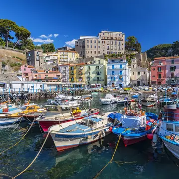 Image of small fishing boats in Sorrento's Marina Grande harbor with colorful buildings and trees in the distance.