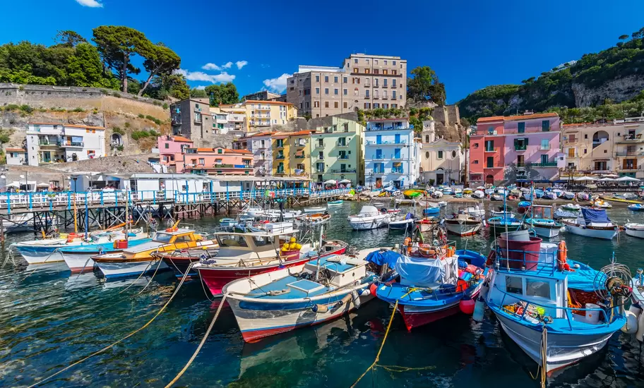 Image of small fishing boats in Sorrento's Marina Grande harbor with colorful buildings and trees in the distance.