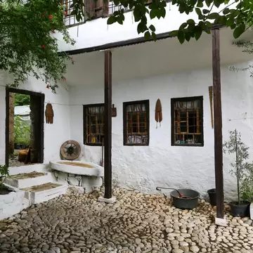A whitewashed Ottoman-era house with dark timber posts, barred windows and a pebbled courtyard shaded by trees.