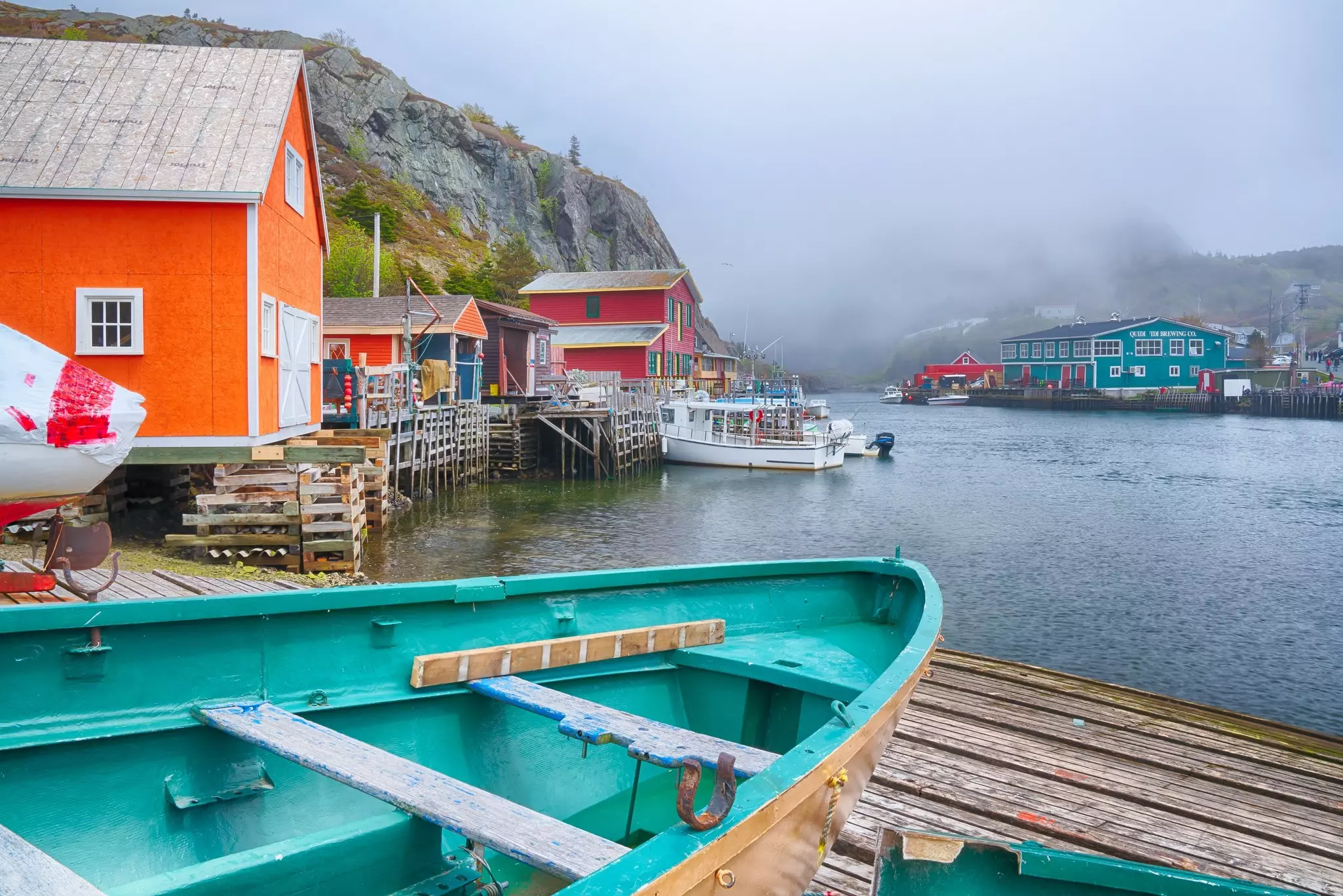 Turquoise-colored rowboat on a dock with colorful fishing buildings in the distance along a harbor on a foggy day.