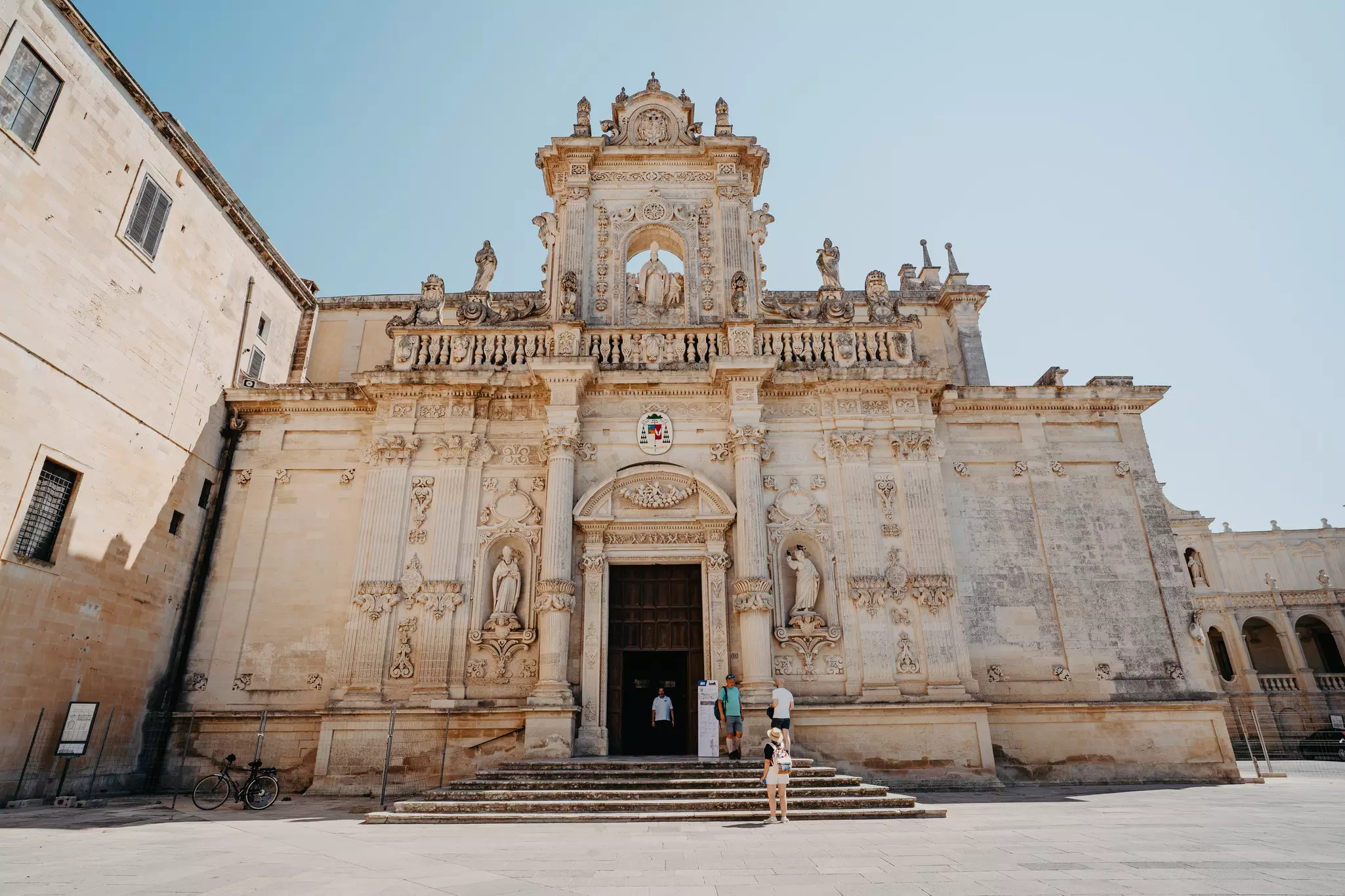 People stand outside a historic cathedral in a city square on a sunny day.