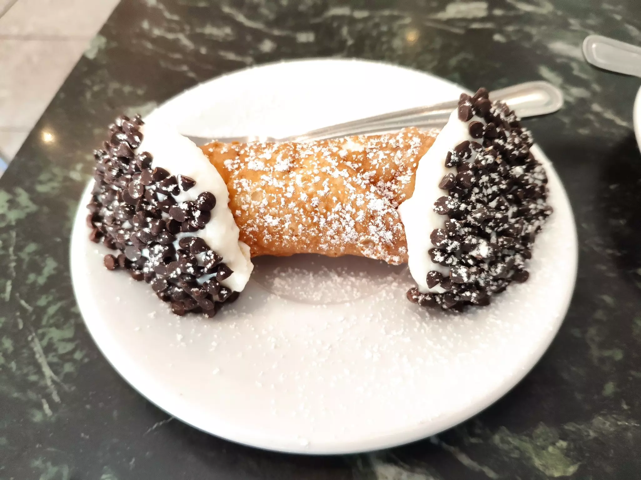 A small plate with a cream-filled pastry on it, with chocolate chips and a dusting of icing sugar.